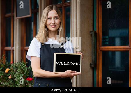 Jeune femme caucasienne de 30 ans avec des cheveux blond courts tenant une pancarte ouverte près des portes du café. Rouvrir le restaurant après le coronavirus Covid-19 Banque D'Images