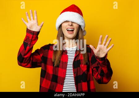 Jeune femme avec chapeau de père noël et chemise à carreaux rouges sur les yeux essayant de saisir quelque chose avec les mains sur jaune arrière-plan Banque D'Images