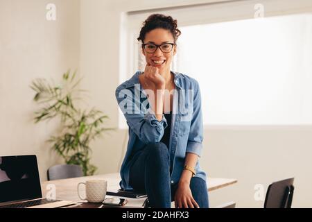 Portrait d'une jeune femme heureuse assise sur une table avec une tasse de café et un ordinateur portable. Femme assise au bureau à domicile. Banque D'Images