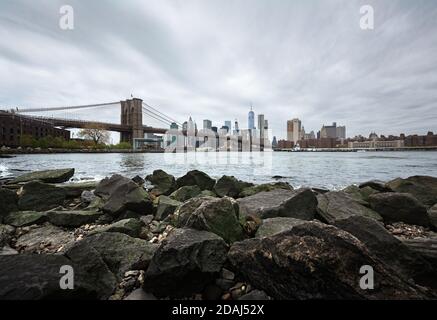 NEW YORK, États-Unis - 29 avril 2016 : gratte-ciel de Manhattan avec pont de Brooklyn. Rochers et pierres sur la rive de la rivière est. Horizon de Manhattan depuis Dumbo Banque D'Images