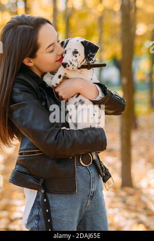 Une jeune femme tient et embrasse un chiot de chien dalmatien dans ses mains tout en marchant dans le parc d'automne. Banque D'Images