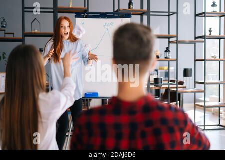 REDHEAD Business Woman finissant le rapport sur le tableau. Des collègues mécontents lancent des boules de papier froissés Banque D'Images