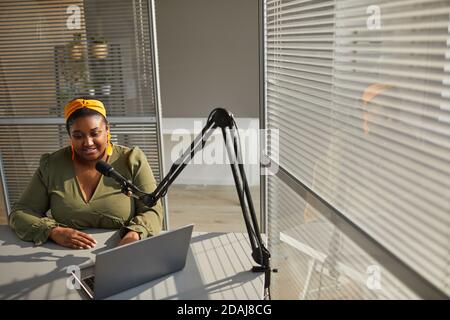 Femme heureuse africaine assise à la table et dactylographiant dessus ordinateur portable lors de la diffusion en direct au bureau Banque D'Images