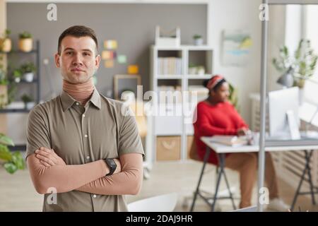 Portrait du jeune chef d'entreprise debout avec les bras croisés et regarder la caméra au bureau avec une femme travaillant dans le arrière-plan Banque D'Images