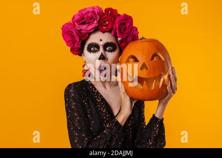 Une jeune fille choquée dans le maquillage d'halloween posant avec la citrouille isolée sur fond jaune Banque D'Images