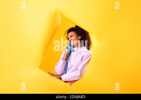 bonne femme afro-américaine parlant sur un téléphone rétro près du trou dans du papier déchiré sur fond jaune Banque D'Images