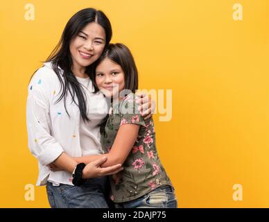 Famille heureuse, maman souriante avec sa fille. Jeune femme et jeune fille vêtue de vêtements décontractés pose embrassant en studio. Isoler sur un jaune Banque D'Images