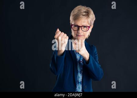 Femme d'affaires blonde plus âgée veut vous combattre en portant des lunettes et une veste bleue Banque D'Images