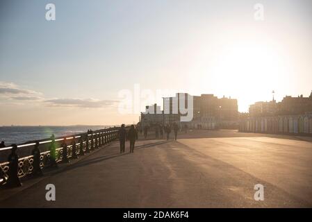 Hove Promenade par Hove pelouses et plage huttes tandis que le soleil se couche derrière les bâtiments. On peut voir des gens marcher le long de l'autre. Le ciel donne une lueur dorée. Banque D'Images