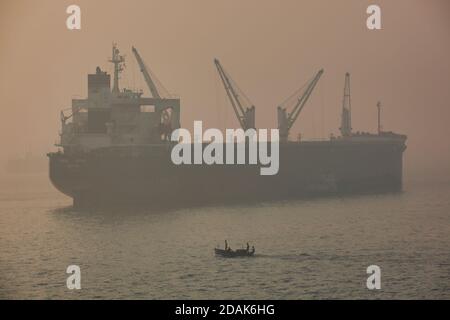 A small motor boat makes it's way across the sea off Mumbai harbour with a cargo ship in the background seen through an early morning haze, India Stock Photo