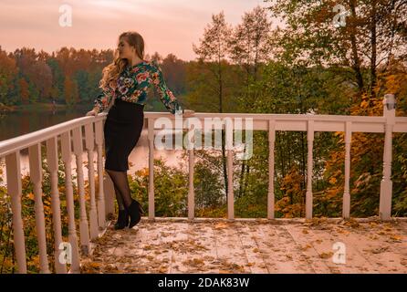 Un adolescent souriant se dresse sur une balustrade blanche qui regarde la caméra blonde, dans une chemise colorée ayant un salon d'automne innocent sur le fond de Banque D'Images