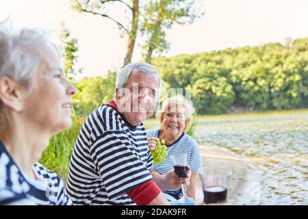 Groupe d'aînés heureux faisant pique-nique au bord du lac et buvant verre de vin rouge Banque D'Images