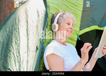 Femme âgée avec une tablette et un casque devant la tente dans la nature en été Banque D'Images