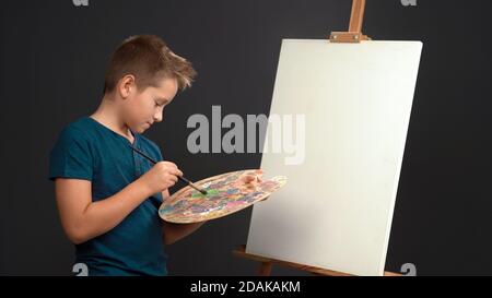 Jeune peintre créatif adolescent garçon mélange des peintures à l'huile sur la palette tout en se tenant devant une toile vierge sur le chevalet. Concept de dessin. Découpez sur le gris Banque D'Images