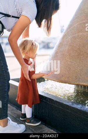 Un enfant mignon joue près de la fontaine avec elle mère un jour ensoleillé Banque D'Images