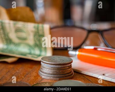 Pile de pièces, floue pliée un billet de dollar américain, un bloc-notes avec un stylo et des lunettes en arrière-plan et une pile de pièces au premier plan. Banque D'Images