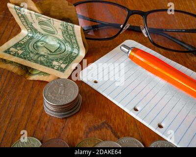 Plié un billet de banque en dollars US, pile de pièces (un quart de dollar US sur le dessus), un bloc-notes avec un stylo, des lunettes et une pile de pièces au premier plan. Banque D'Images