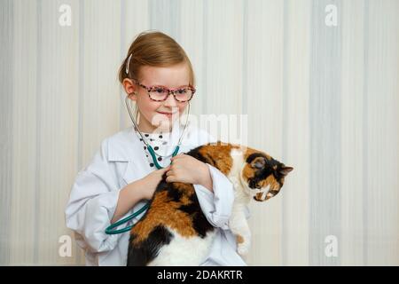 Une petite fille dans un manteau de docteur sourit et invite à un traitement. Le patient est un chat domestique. Jeu médical éducatif. Banque D'Images