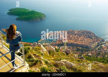 Attrayant voyageur de brunette assis sur une rampe au-dessus de la ville de Dubrovnik, point de vue sur la montagne SRD. Vue sur l'ancienne ville historique surrou Banque D'Images