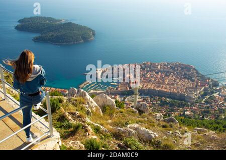Attrayant voyageur de brunette assis sur une rampe au-dessus de la ville de Dubrovnik, point de vue sur la montagne SRD. Vue sur l'ancienne ville historique surrou Banque D'Images