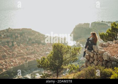 Gros plan d'une brunette assise sur le bord d'un rocher posant pendant un coucher de soleil lumineux avec la vieille ville de Dubrovnik derrière elle au loin. Visitin Banque D'Images