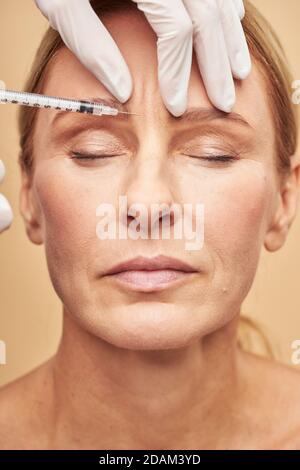 Gros plan des mains de l'esthéticienne dans des gants de protection faisant l'injection sur le front féminin Banque D'Images