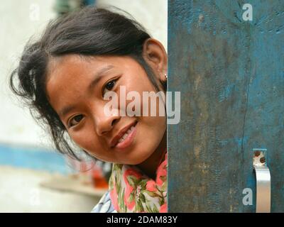 Jolie fille indienne du nord-est Apatani sourit pour l'appareil photo et peeks autour d'une porte en bois bleu. Banque D'Images