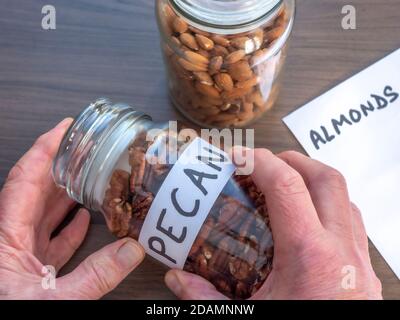 Gros plan POV des mains d'un homme collant une étiquette écrite pour noix de pécan sur un pot en verre plein, avec un autre pot contenant des amandes en arrière-plan. Banque D'Images