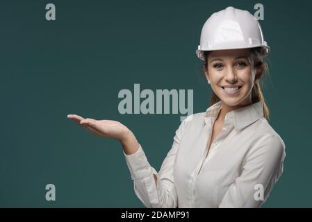 Jeune ingénieur souriant portant un casque de sécurité et présentant quelque chose avec sa main Banque D'Images