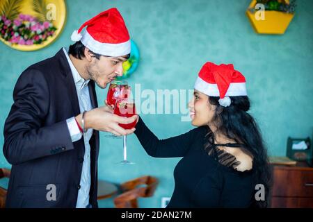 Joyeux jeune couple indien en chapeaux de père noël célébrant ensemble Noël tout en s'amusant avec des verres à cocktail rouge amour, collage, Banque D'Images