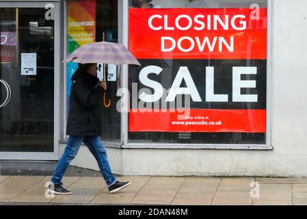 Bridport, Dorset, Royaume-Uni. 14 novembre 2020. Maintien Covid-19. Une femme avec un parapluie passe devant une branche de paons à Bridport dans Dorset qui a une fermeture de l'affiche solde dans la fenêtre du magasin lors d'une matinée humide pendant le deuxième confinement de Covid-19. Crédit photo : Graham Hunt/Alamy Live News Banque D'Images