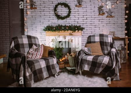 Deux fauteuils couverts de couvertures confortables debout près de la cheminée décorée de guirlande, bougies et panier avec des cadeaux. Couronne ronde verte suspendue Banque D'Images