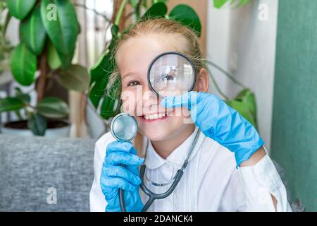 Une fille de 9-10 ans de race blanche, revêtant un manteau blanc, des gants médicaux bleus tient un stéthoscope dans sa main, regarde à travers une loupe, sourit. Rés Banque D'Images