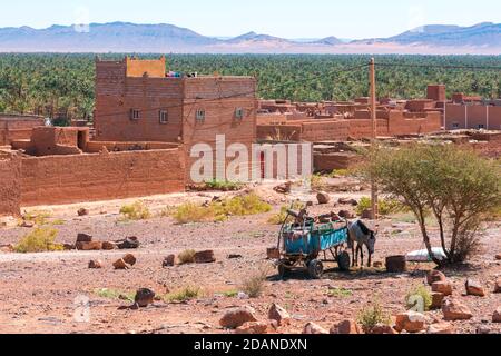 Âne avec charrette et maisons en adobe en premier plan et forêt de palmiers et montagnes de la vallée de Draa, Zagora, Maroc. Banque D'Images