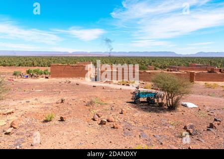 Âne avec charrette et maisons en adobe en premier plan et forêt de palmiers et montagnes de la vallée de Draa, Zagora, Maroc. Le feu de camp fume dans l'arrière-pays Banque D'Images