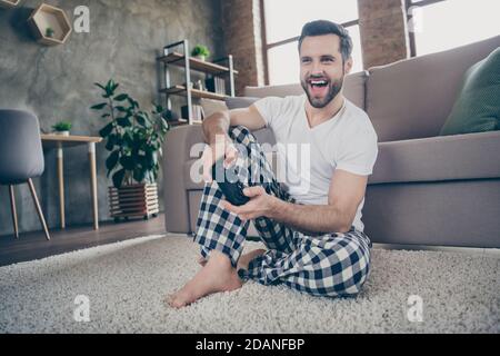 Photo de tout le corps du contenu Guy assis tapis de sol profiter jouer aux jeux vidéo joystick porter damier pantalon pantalons blanc t-shirt à l'intérieur Banque D'Images