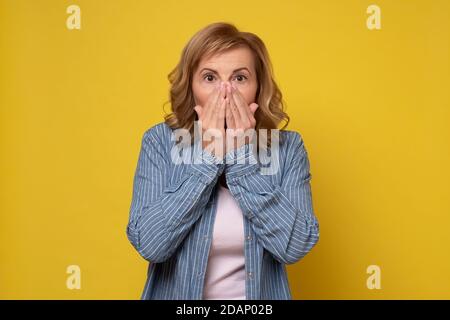 Une femme mature choquée couvre sa bouche ouverte avec la main, surprise par les nouvelles. Banque D'Images