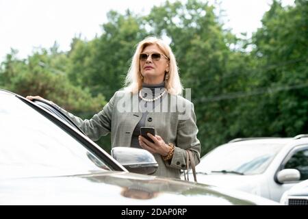 Une femme d'affaires élégante et mature utilisant un smartphone en restant debout voiture Banque D'Images