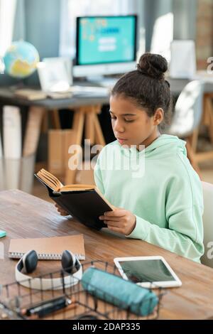 Mignonne école élémentaire dans vêtements décontractés lecture livre par table dans séjour Banque D'Images