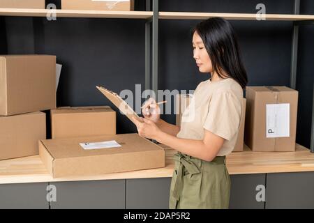 Jeune femme avec stylo et presse-papiers vérifiant les ordres de clients de la boutique en ligne Banque D'Images