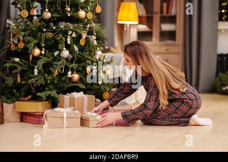 Blonde jeune femme en pyjama mettant des cadeaux de Noël sous décoré firtree Banque D'Images