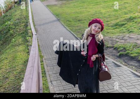 Une dame souriante se promette dans les escaliers et porte une palette bordeaux et le biret, souriant à l'appareil photo dans des vêtements noirs, à l'automne contre le Banque D'Images