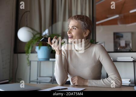 Femme d'affaires souriante enregistrant un message vocal sur son téléphone au bureau Banque D'Images