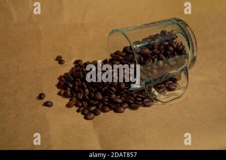 Grains de café torréfiés déversés d'une tasse en verre transparent sur fond marron Banque D'Images