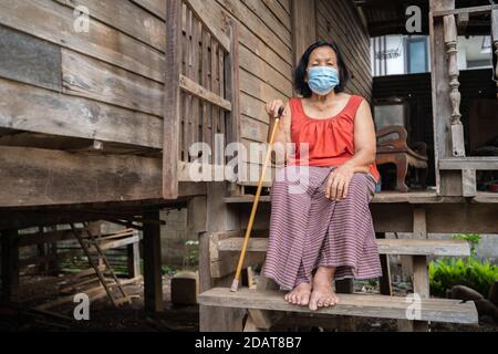 Femme thaïlandaise âgée en col rond sans manches portant un masque médical pour protéger la pandémie du virus corona (covid-19) dans une ancienne maison en bois Banque D'Images