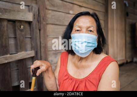 Femme thaïlandaise âgée en col rond sans manches portant un masque médical pour protéger la pandémie du virus corona (covid-19) dans une ancienne maison en bois Banque D'Images