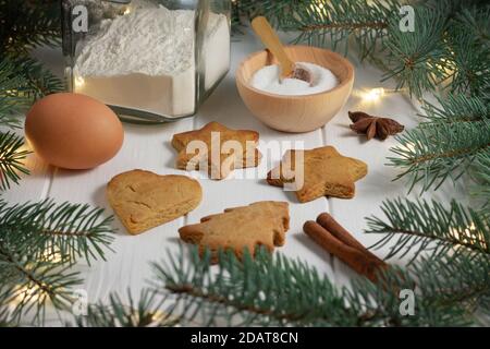 Pain d'épice, farine, œuf, sucre, anis, cannelle à côté des branches vertes d'un arbre de Noël avec guirlande lumineuse LED sur fond de bois blanc. Banque D'Images