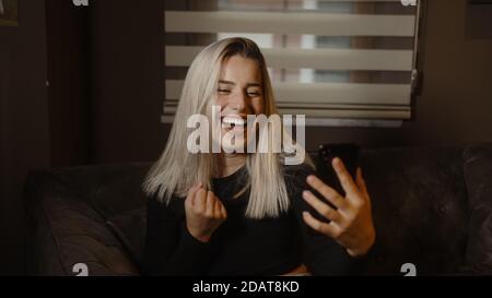 Jeune femme blonde douce assise sur un canapé et criant de joie à la fois tout en regardant son téléphone, la technologie de style de vie et le concept de shopping Banque D'Images