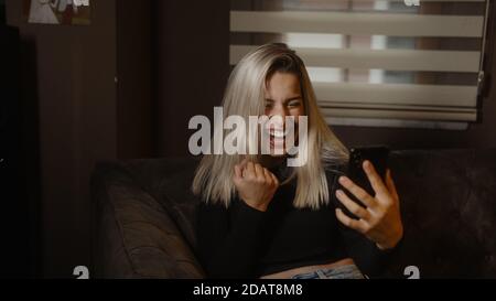 Jeune femme blonde douce assise sur un canapé et criant de joie à la fois tout en regardant son téléphone, la technologie de style de vie et le concept de shopping Banque D'Images