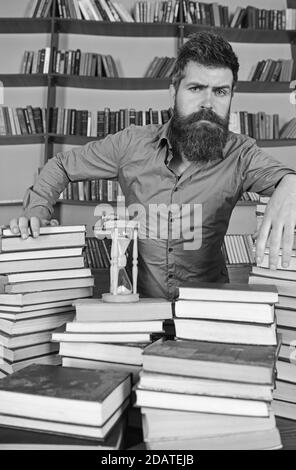 Concept de bibliothécaire. Professeur, scientifique avec la barbe se tient à table avec des livres, défoqué. L'homme sur le visage réfléchi se tient entre des piles de livres, tout en étudiant dans la bibliothèque, les étagères sur le fond. Banque D'Images